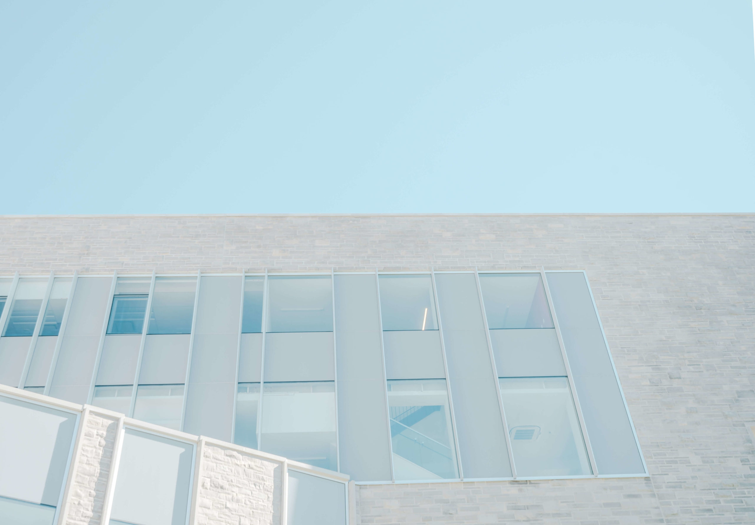 Modern building facade with large rectangular windows, showcasing a minimalist and clean design against a clear blue sky. The structure features light-colored brick and glass elements.