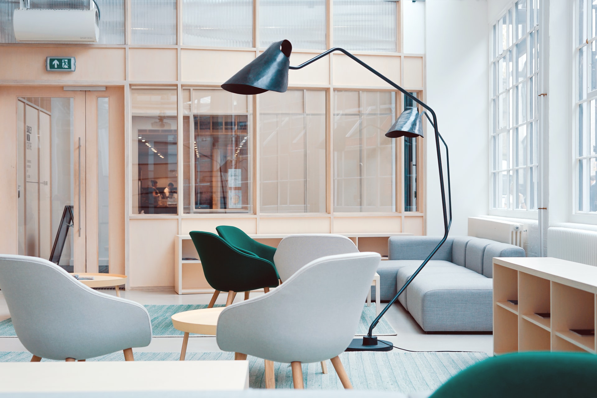 A modern lounge area with light blue and green armchairs, a gray sofa, and a large black floor lamp. Wooden tables and a light blue rug complement the space, which is enclosed by large windows and wooden paneling.