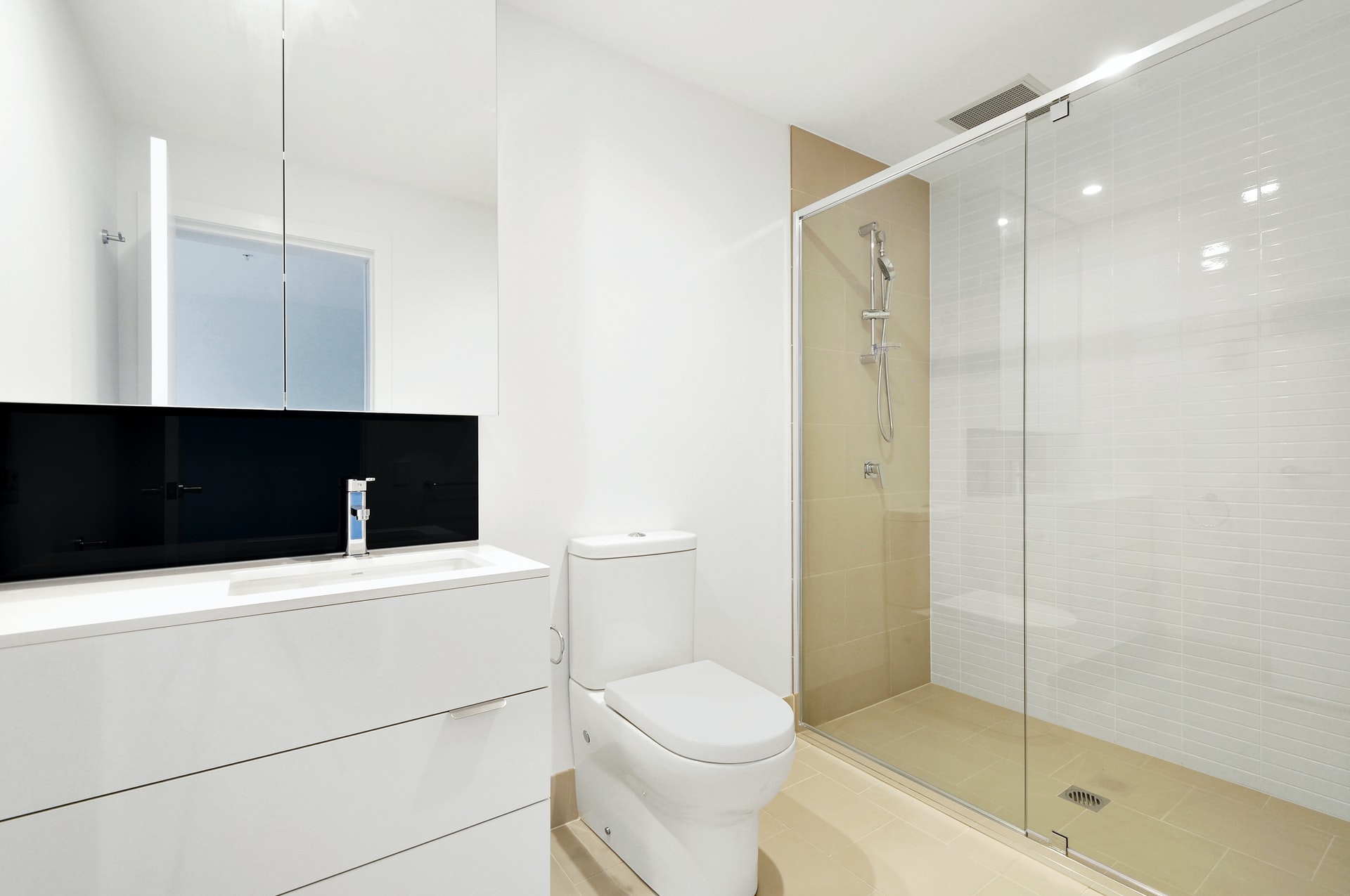 A modern bathroom with a white sink and vanity on the left, a toilet in the center, and a glass-enclosed shower with a chrome showerhead on the right. Walls are white, and the floor is tiled in beige.