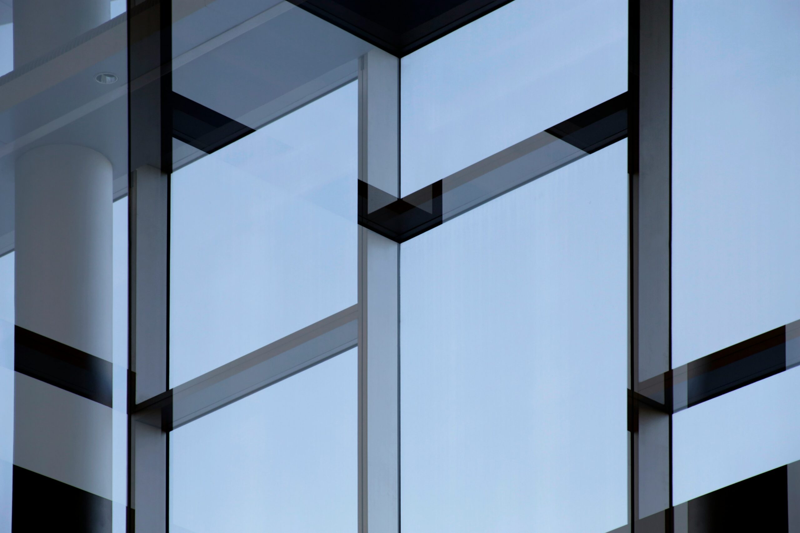 Modern building facade featuring large, reflective glass panels arranged in geometric patterns with visible black frames. The blue sky is faintly reflected in the windows, creating a sleek and minimalist appearance.