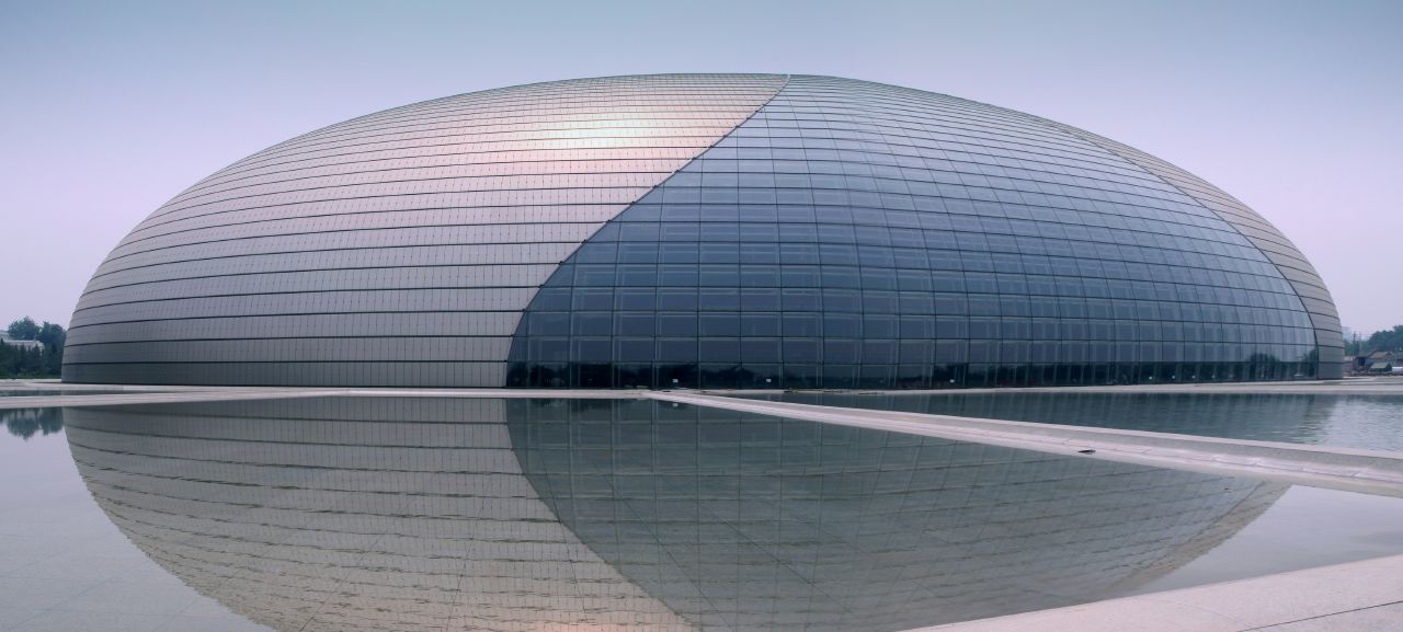 The image shows the National Centre for the Performing Arts in Beijing, China. Its modern, dome-shaped structure is made of glass and titanium, surrounded by a reflective pool, under a clear sky.