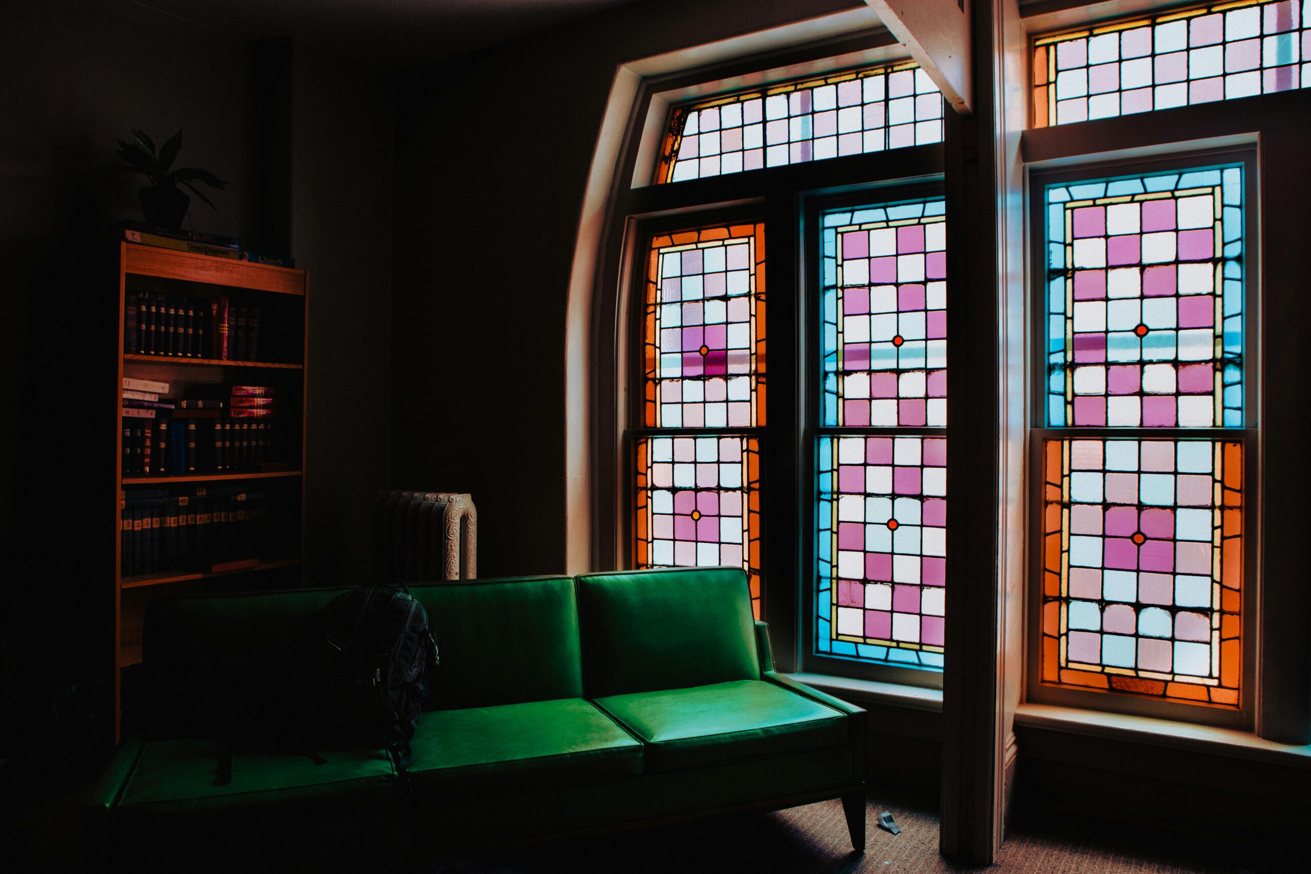 A dimly lit room features a green sofa next to a bookshelf filled with books. Two tall, arched stained glass windows with colorful geometric patterns illuminate the space, creating a vibrant contrast against the dark interior.