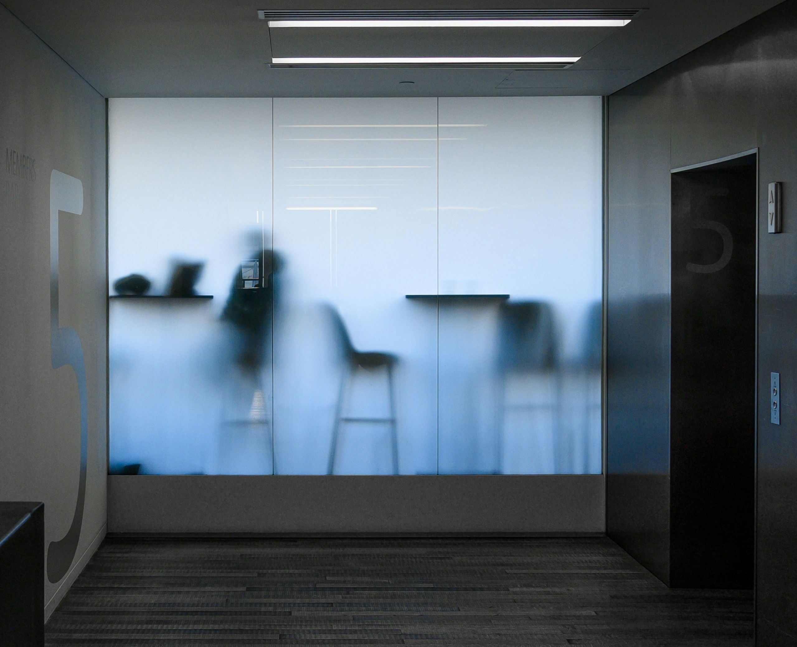 Silhouettes of people sit at high tables behind frosted glass in a modern hallway, with the number 5 marked on the wall and elevator, evoking a calm office or building interior.
