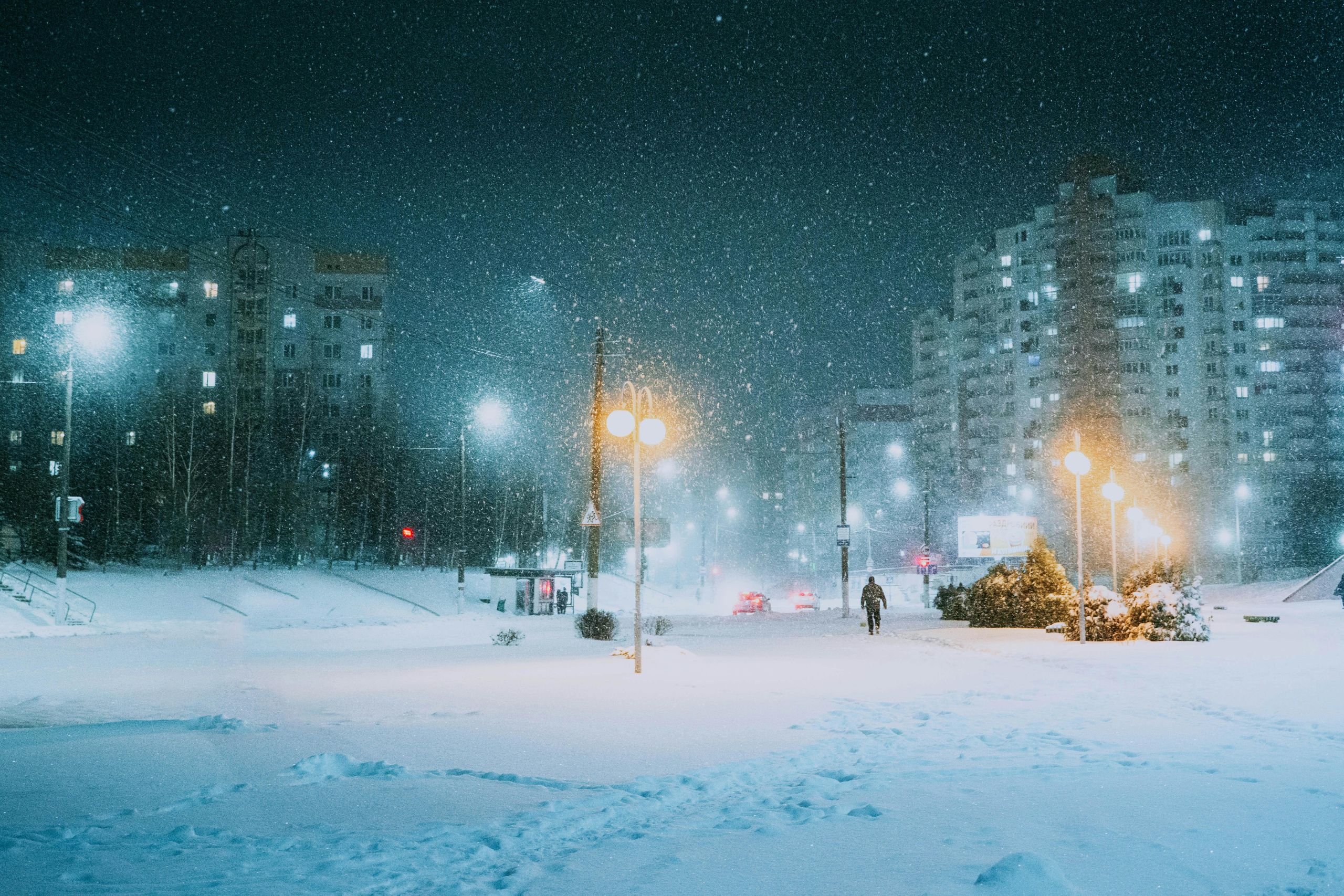 A snowy city street at night is illuminated by streetlights and apartment buildings. Snow falls heavily as a few people and cars pass in the distance—reminding shop owners to protect your storefront glass in this cold, wintry urban scene.