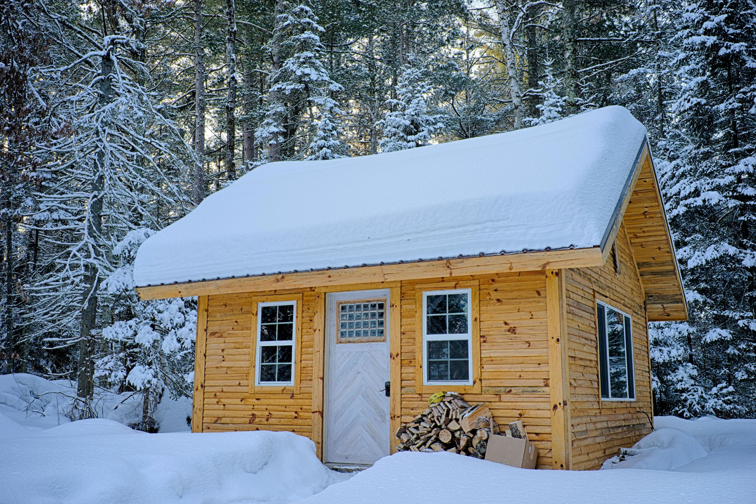 A small wooden cabin with a steep snow-covered roof stands in a snowy forest. Stacked firewood rests by the front door, while tall pine trees surround the cabin—protected from drafts and condensation—under a clear winter sky.