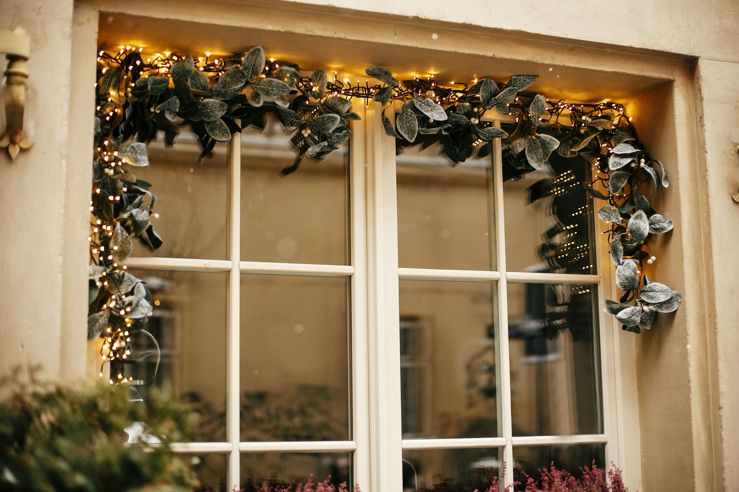 A window, free from common winter window problems, is decorated with frosted green garland and warm white string lights. The window reflects the beige exterior of another building, creating a cozy, festive atmosphere.