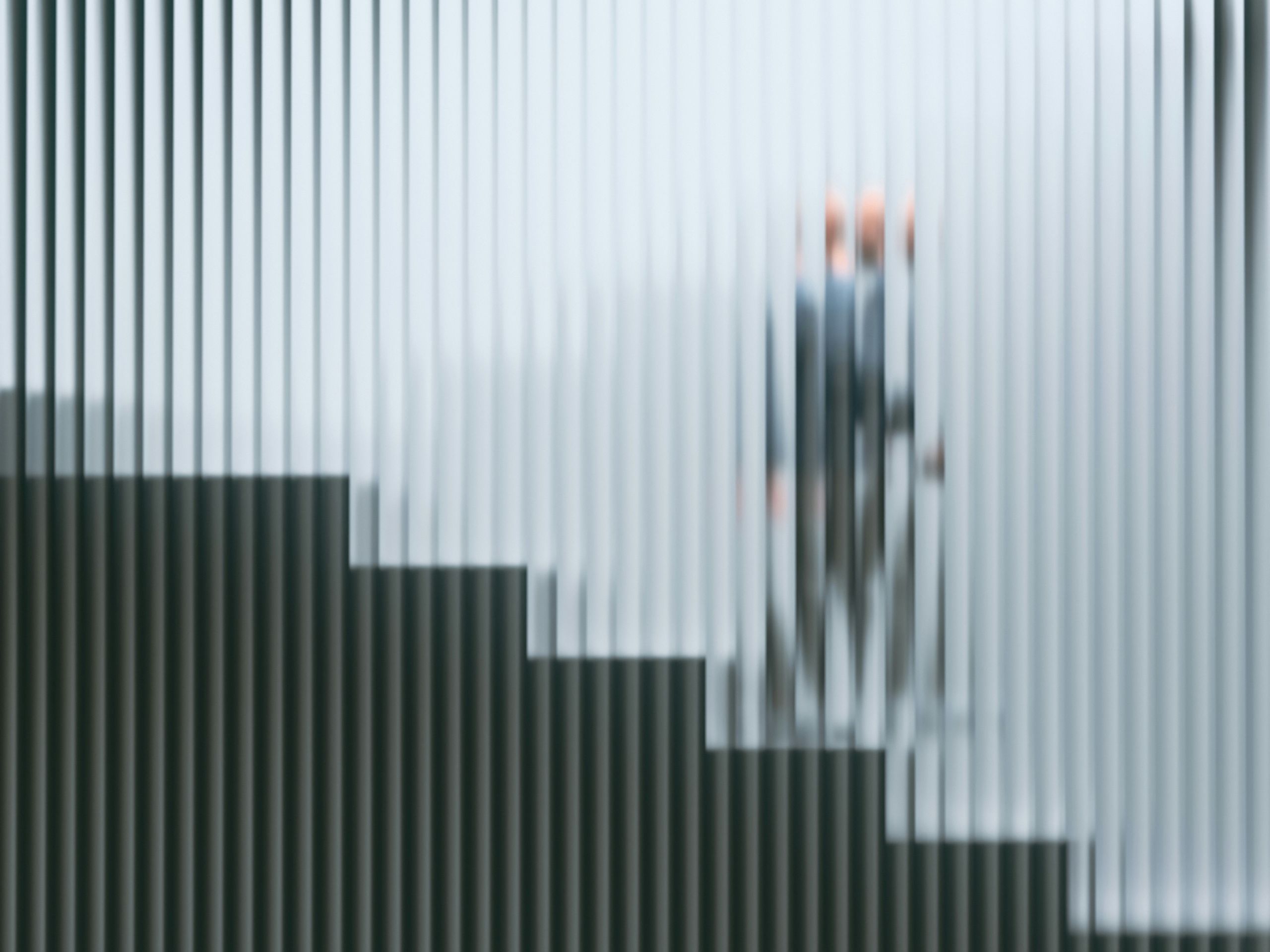 A blurred figure is seen behind custom glass design with vertical stripes, while a staircase stands in the foreground. The image is abstract, obscuring details of the person and creating a modern, geometric effect.