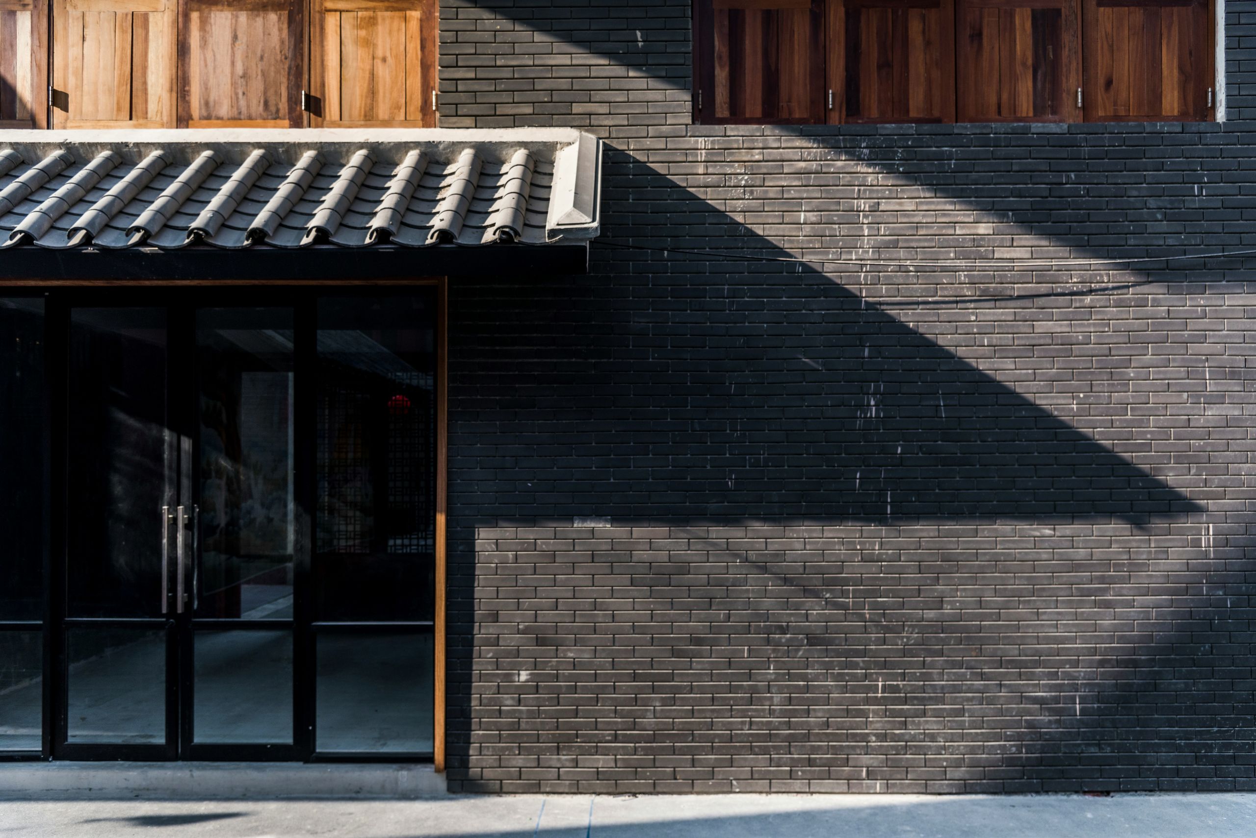 A modern building facade as part of a commercial glass installation, with dark brick walls, large glass double doors on the left, and closed wooden windows above. Sunlight casts sharp geometric shadows across the brick surface.