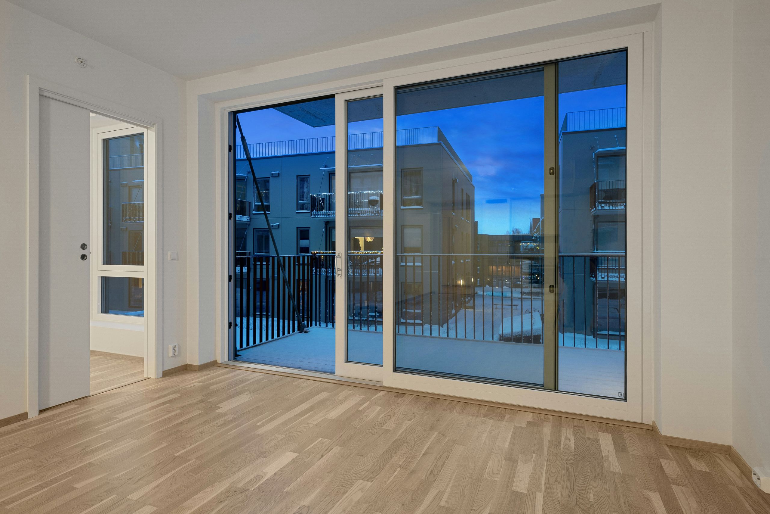 A modern empty room with light wooden floors, large sliding glass doors leading to a balcony, and a view of neighboring apartment buildings at dusk. An open doorway leads to another room—perfect for those considering sliding glass door repair or upgrades.