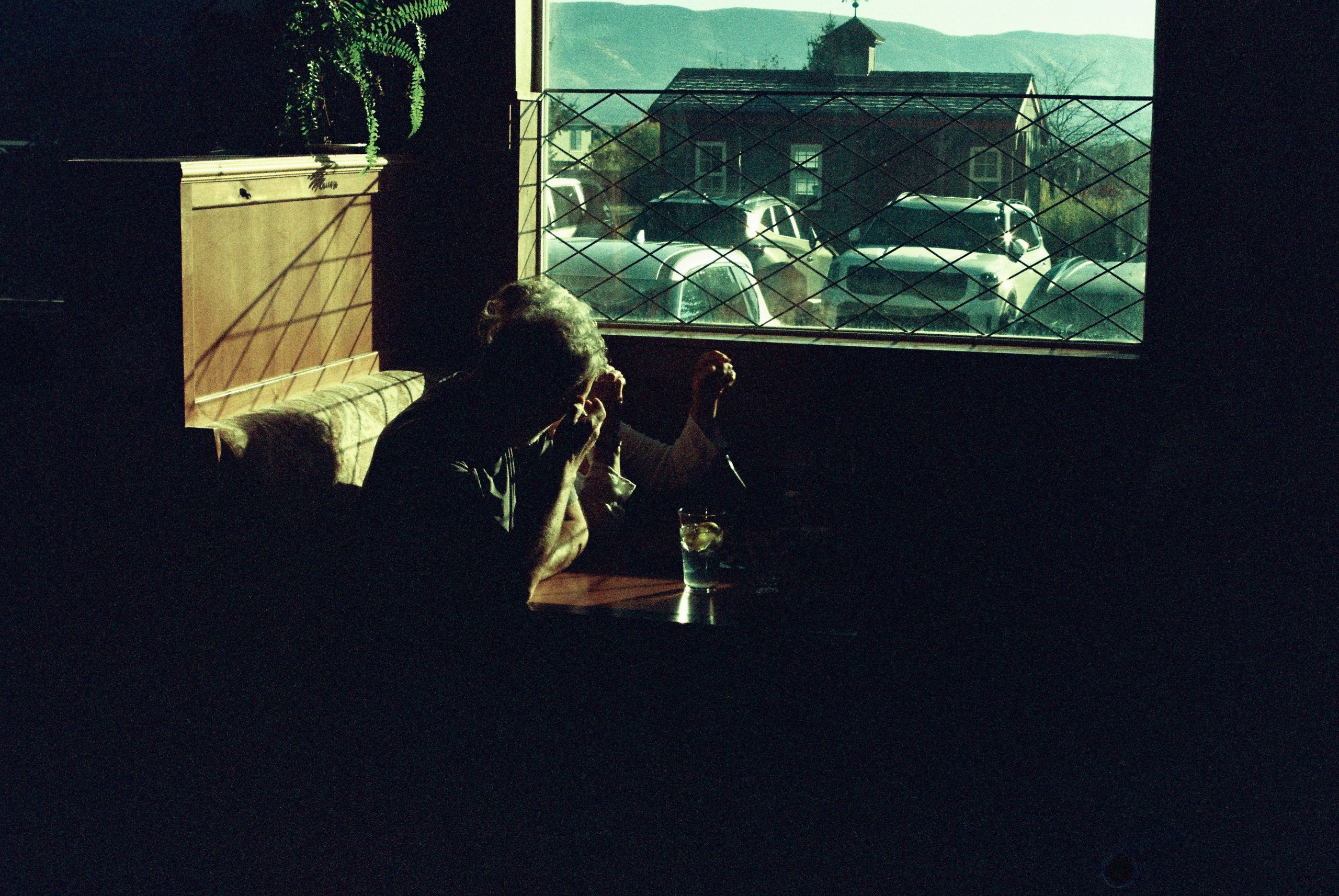 Two people sit at a booth by a sunlit window, its clarity a mark of a local glass company, holding hands in the dim room. Outside, cars and a building shine in daylight. A plant rests on the ledge nearby.