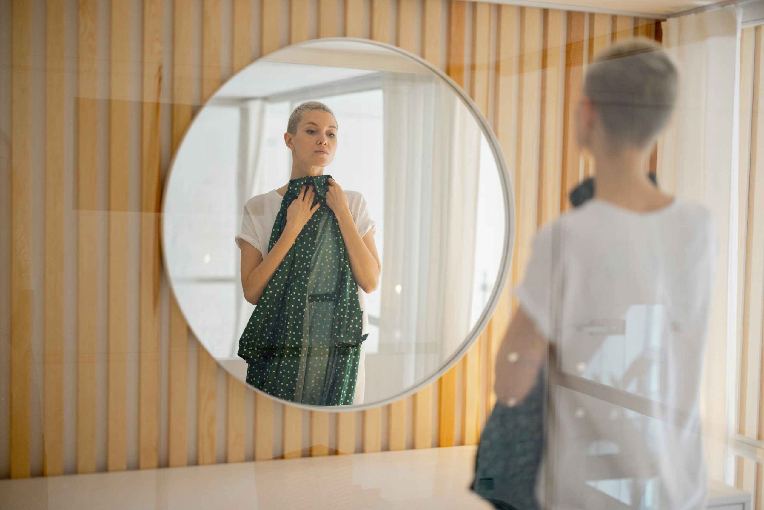 A person with short blond hair looks at themselves in a round custom mirror while holding a dark green, patterned shirt against their chest in a room with wooden wall panels.