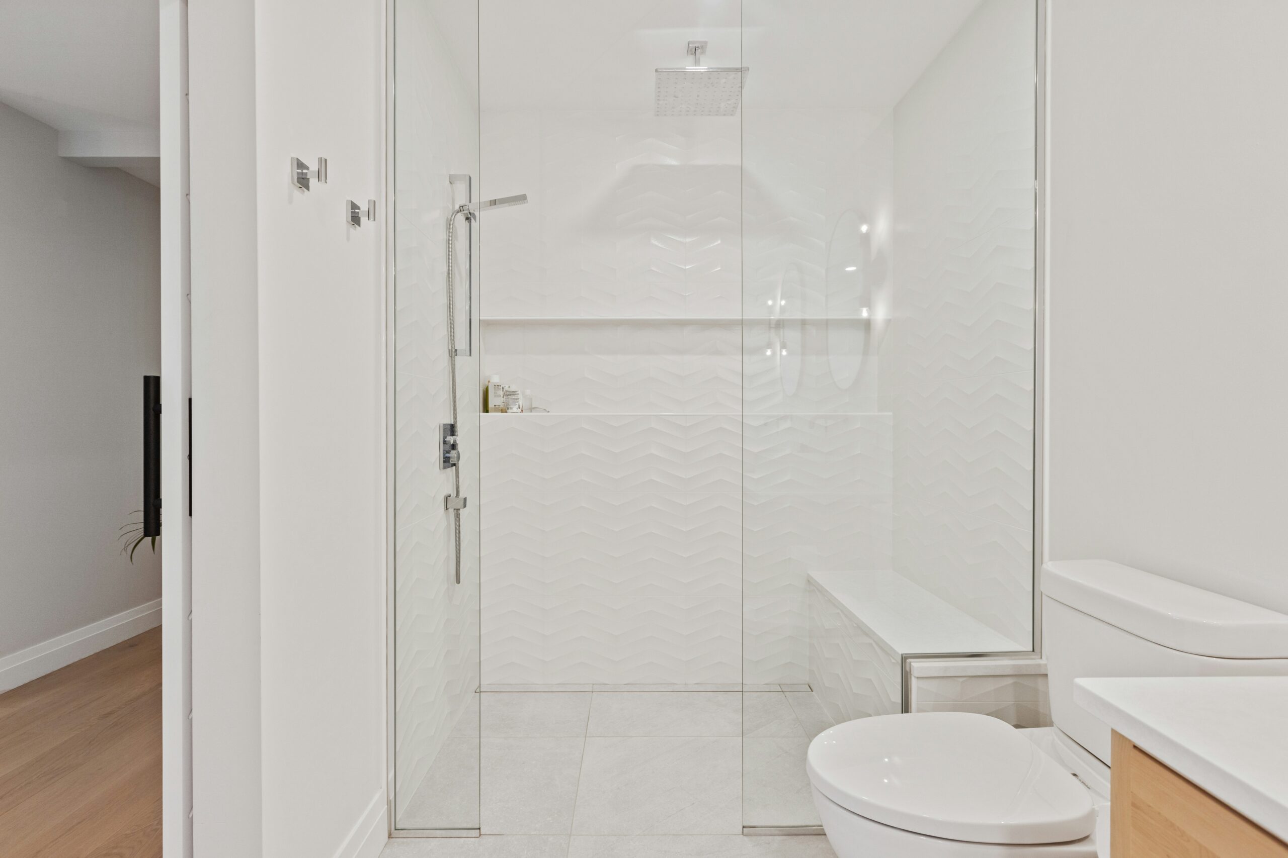 A modern bathroom features spacious framed shower doors enclosing a glass shower, white textured tile walls, a built-in bench, and a rain showerhead. A white toilet sits beside a wooden vanity in the foreground.