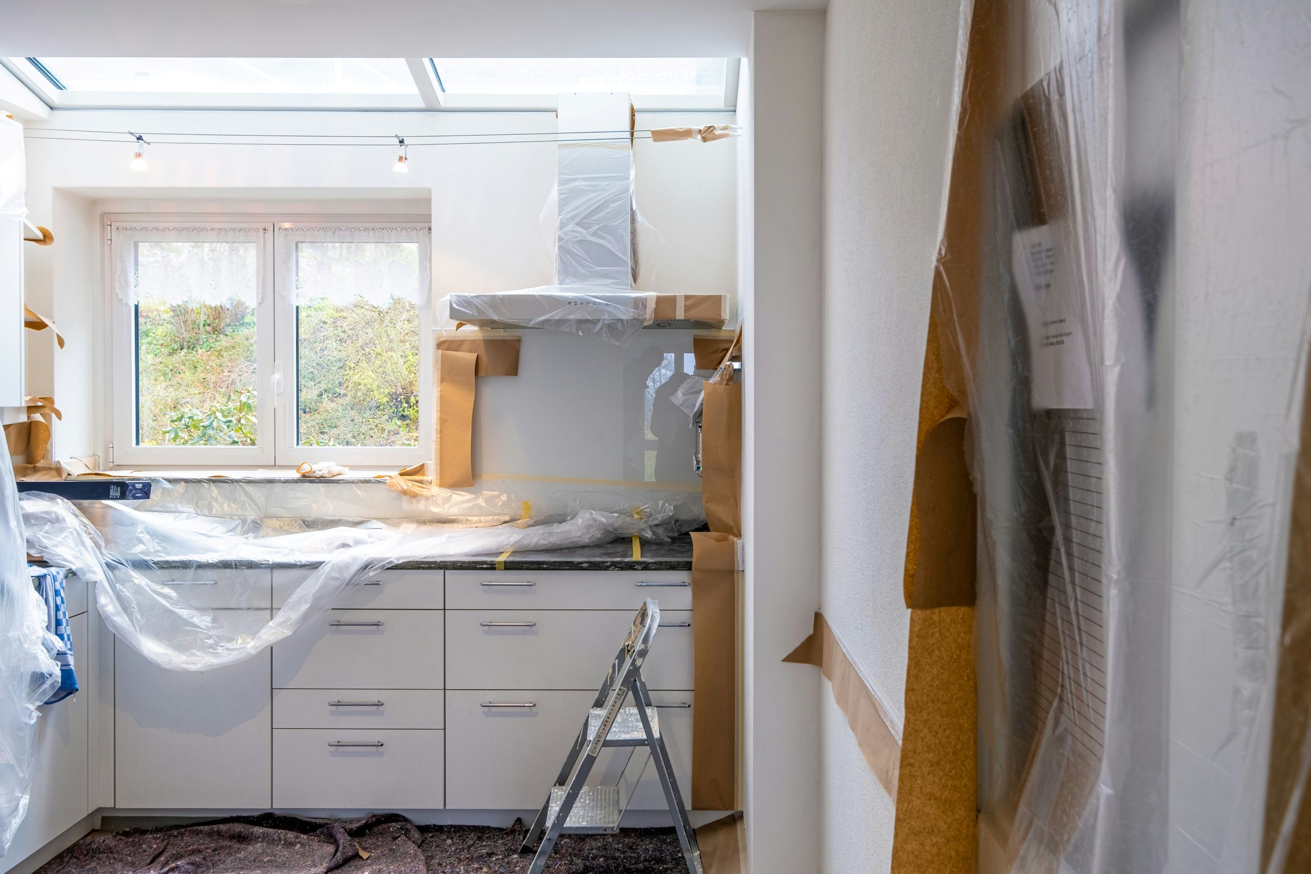 A kitchen under renovation with cabinets and appliances covered in plastic sheets, a step ladder by the counter, and protective materials taped to surfaces. Light from a window above the sink highlights recent work by an Arkansas glass company