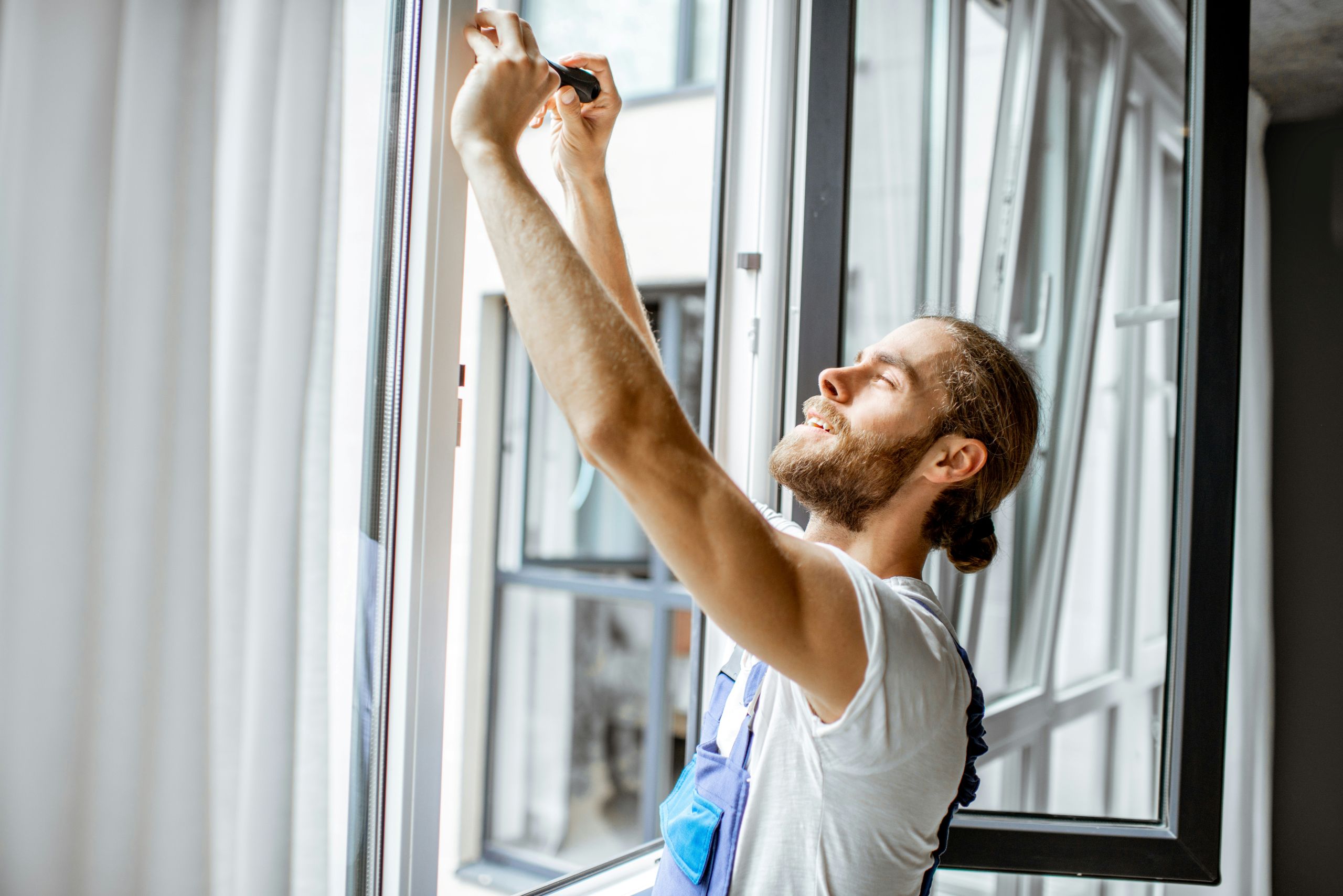A man with a beard and ponytail, wearing overalls, doing a window glass replacement. He installs or adjusts a window screen in a bright room with open windows and sheer white curtains. He is smiling and appears focused on his work.