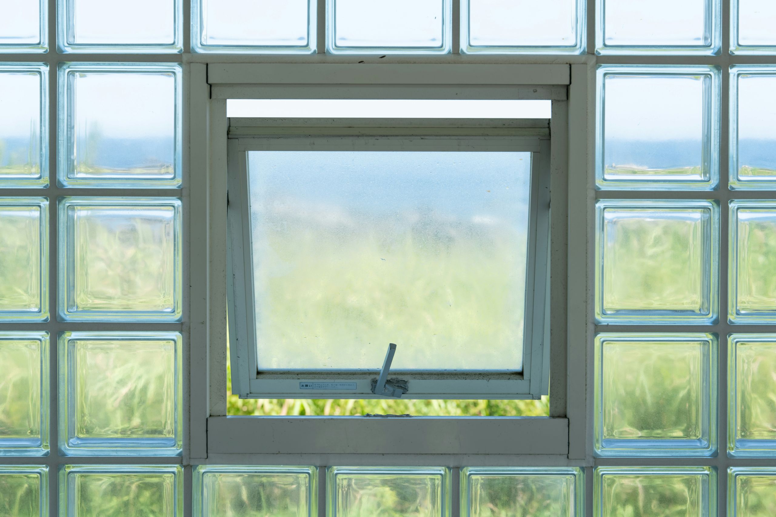 An insulated glass replacement window, slightly open that is centered in a wall of glass blocks. The background shows blurred green foliage.