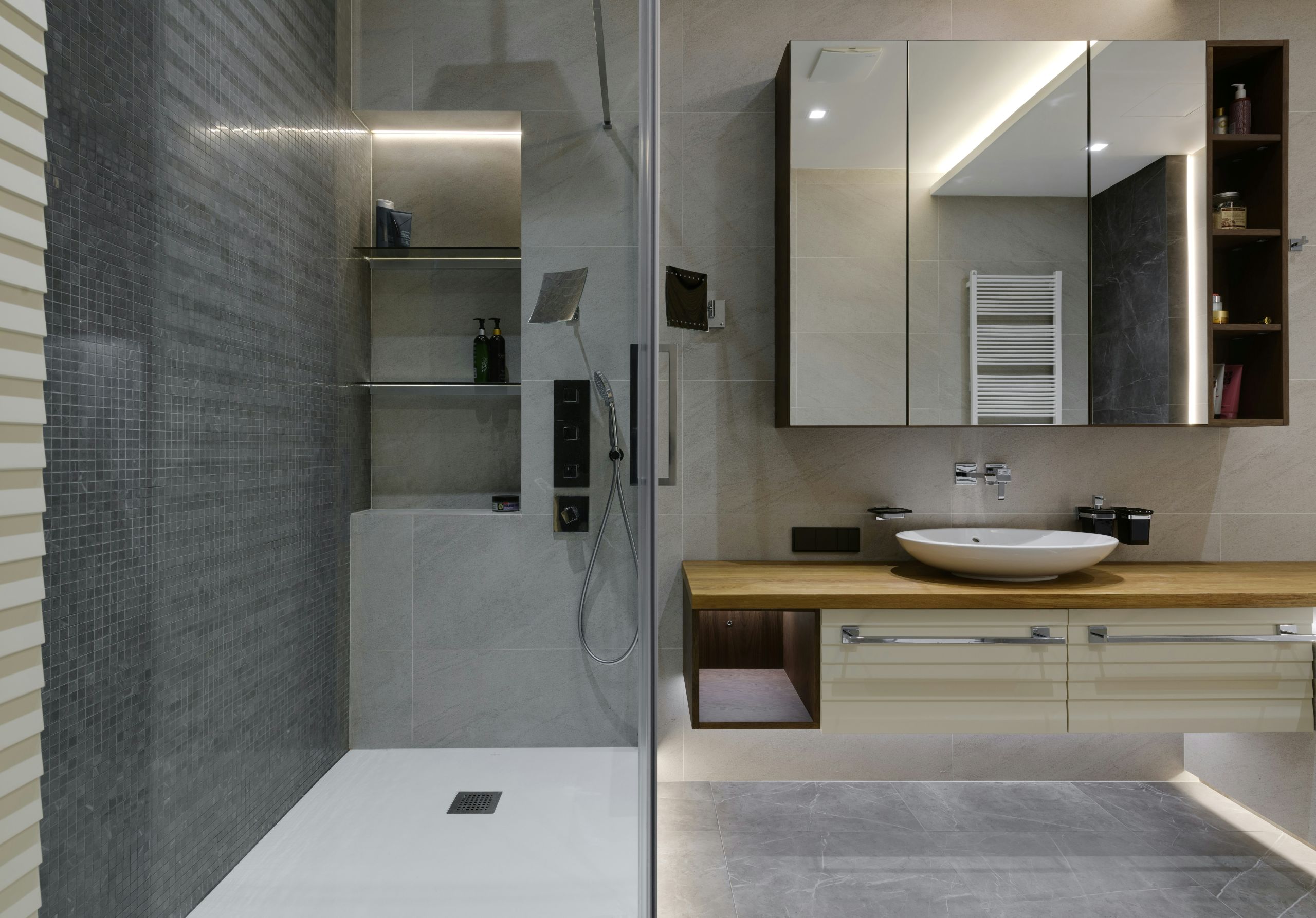 Modern bathroom with a CUSTOM CUT GLASS-enclosed shower, gray tile walls, a wooden vanity with a white vessel sink, large lit mirror, wall shelves, and a towel warmer. The design is sleek and minimalist.