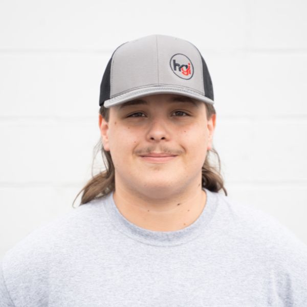 A young person with long brown hair, a faint mustache, and a gray and black baseball cap with an hci logo, wearing a light gray T-shirt, stands in front of a plain white wall.
