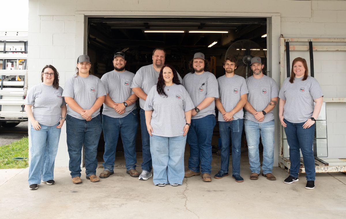 Nine people stand side by side in front of an open garage, all wearing matching gray t-shirts and jeans, smiling at the camera. The background shows tools and equipment inside the garage.