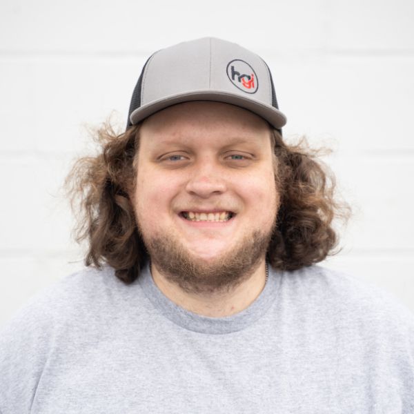A man with curly brown hair and a beard smiles at the camera. He is wearing a gray and black baseball cap with a logo, and a light gray T-shirt. The background is a plain white wall.