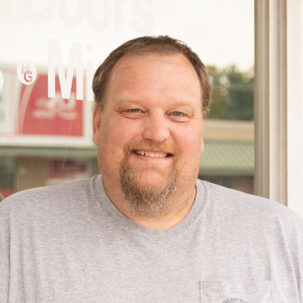 A man with light brown hair and a beard is smiling at the camera. He is wearing a light gray T-shirt and standing in front of a window with faint white text. The background shows part of a building and trees.