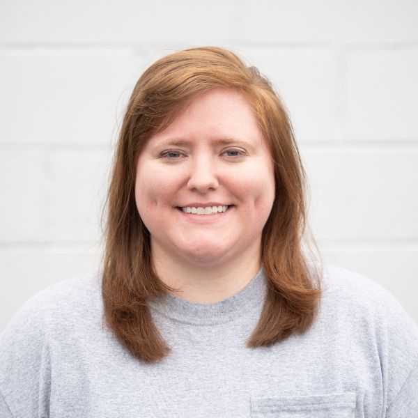 A person with light skin and straight, shoulder-length auburn hair is smiling while standing in front of a white brick wall. They are wearing a light gray crew-neck shirt.