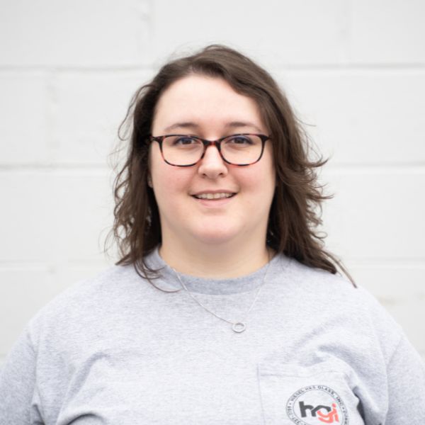 A person with shoulder-length brown hair and glasses, wearing a gray shirt with a small logo, stands in front of a light-colored brick wall, smiling at the camera.