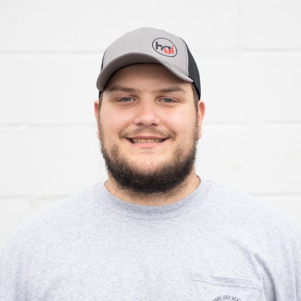 A man with a beard wearing a gray T-shirt and a gray and black baseball cap stands in front of a white wall, smiling at the camera.