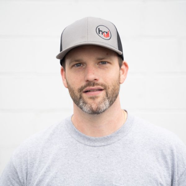 A man with a short brown beard and mustache wears a gray and black cap with a circular logo, and a light gray T-shirt, standing against a plain white background.