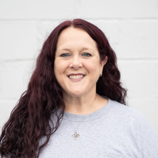 A woman with long, wavy dark red hair smiles at the camera. She is wearing a light gray top and a silver necklace with a cross pendant. The background is a plain white wall.