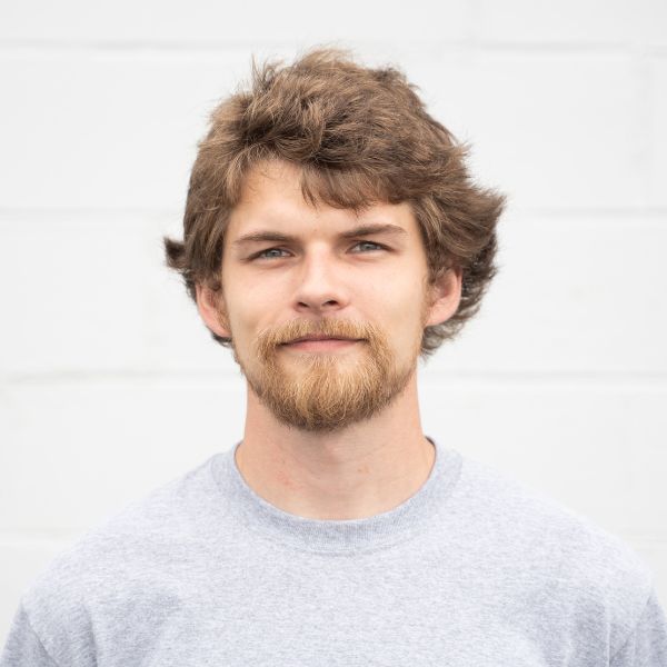 A young man with wavy brown hair, a beard, and mustache stands in front of a white brick wall, wearing a light gray crew-neck shirt and looking directly at the camera with a neutral expression.