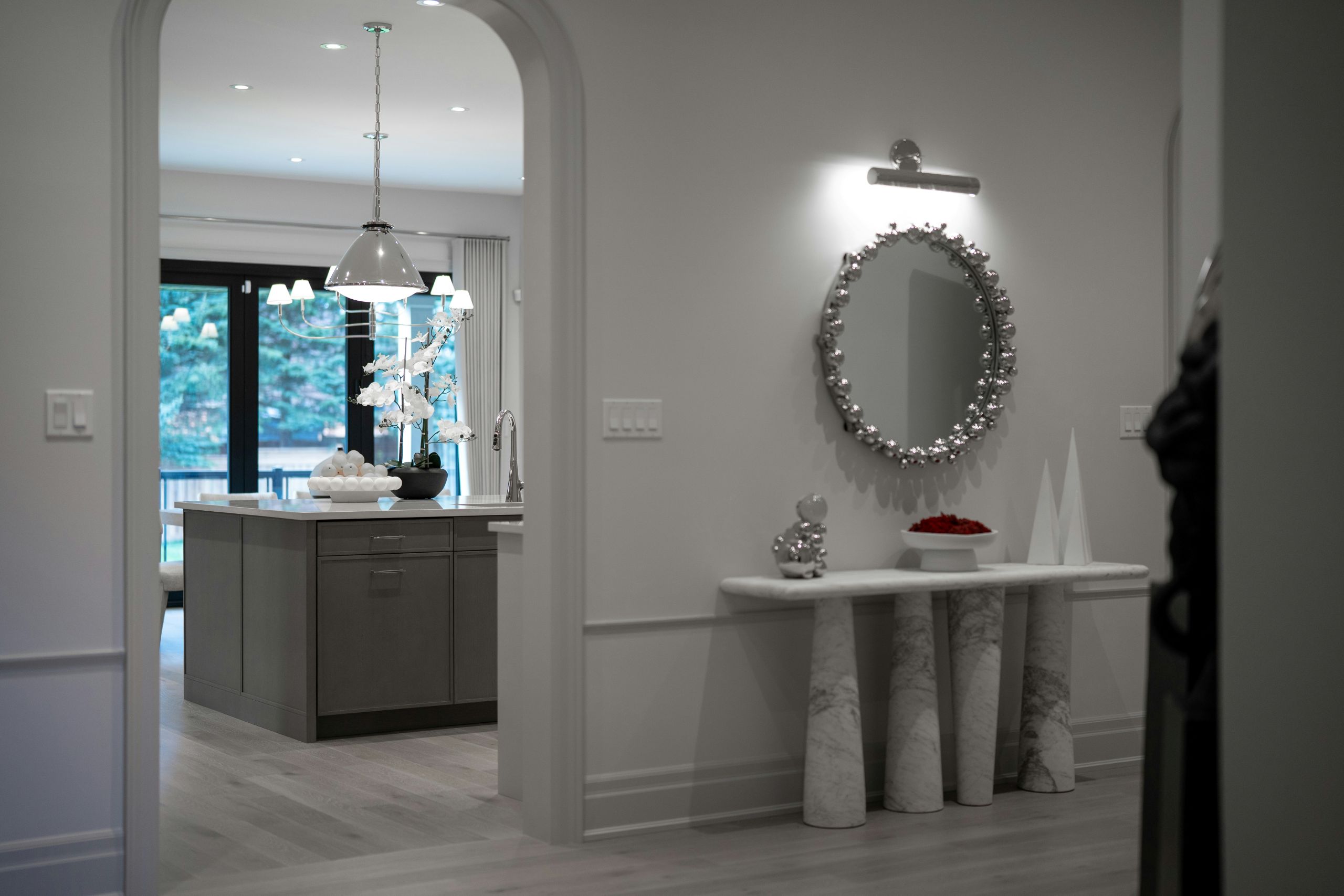 A modern hallway with a marble console table, round decorative mirror, and minimalist decor leads to a bright kitchen featuring an island, pendant lights, and large windows with recent glass replacement overlooking greenery.