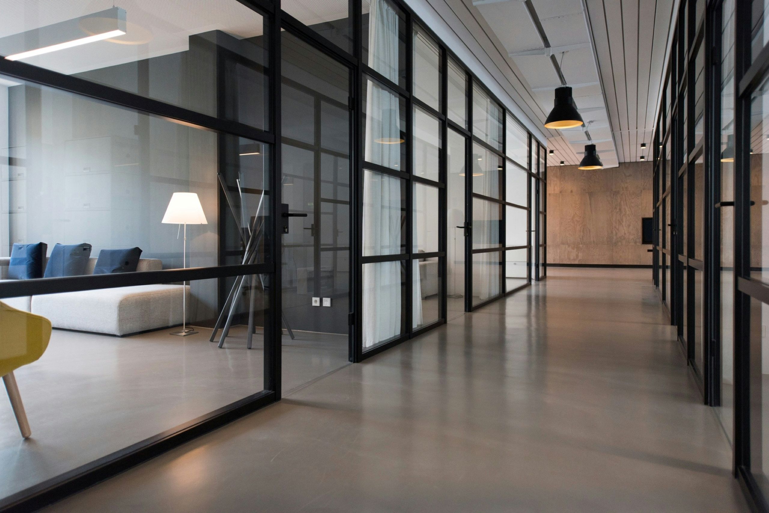 a conference room glass in an office hallway, a gray sofa, floor lamp, yellow chair, and black ceiling lights. The space is minimalistic with polished concrete floors and a neutral color palette.