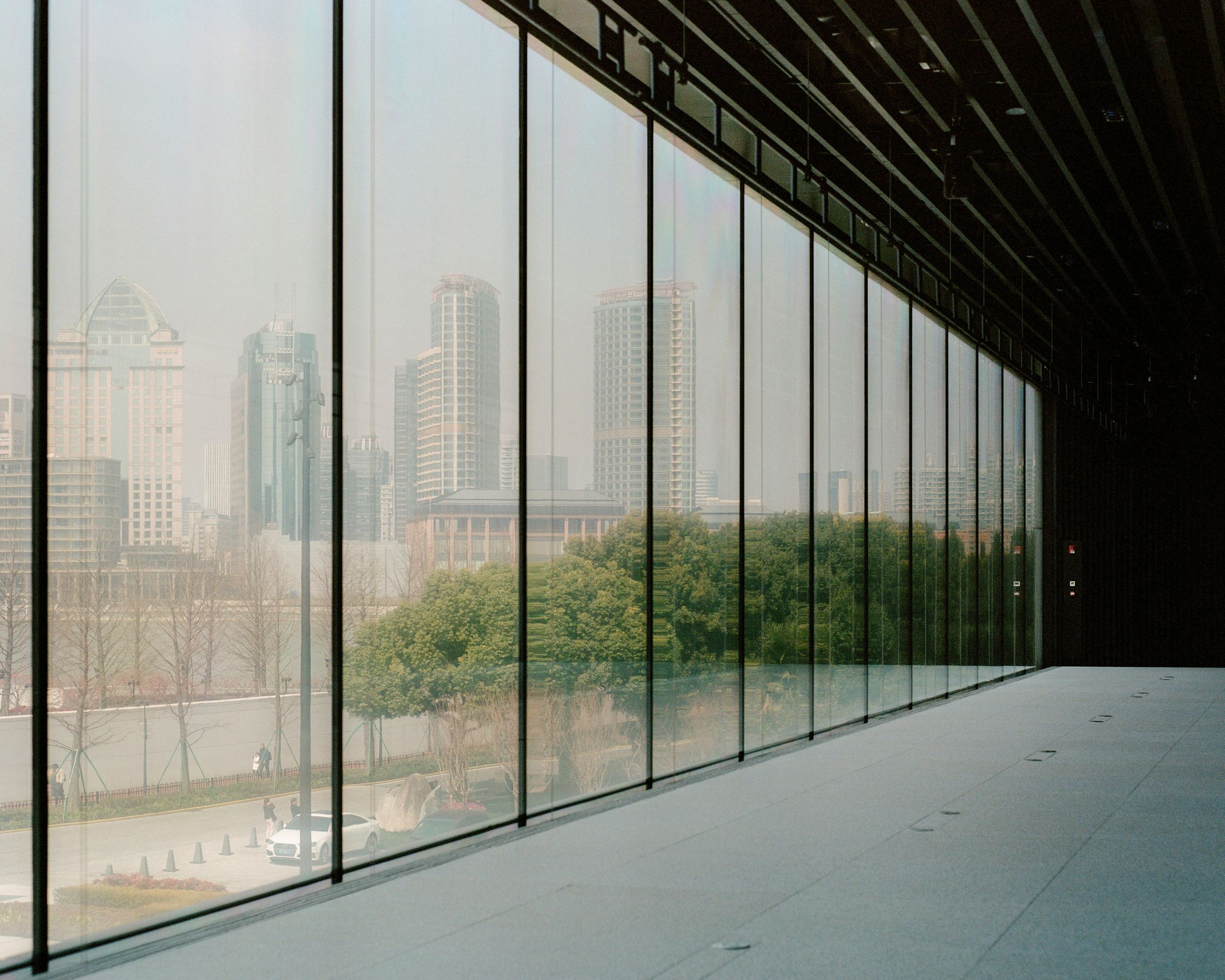 A modern cityscape showing storm window protection. It shows tall buildings seen through large floor-to-ceiling glass windows, reflecting trees and a white car outside on a cloudy day.