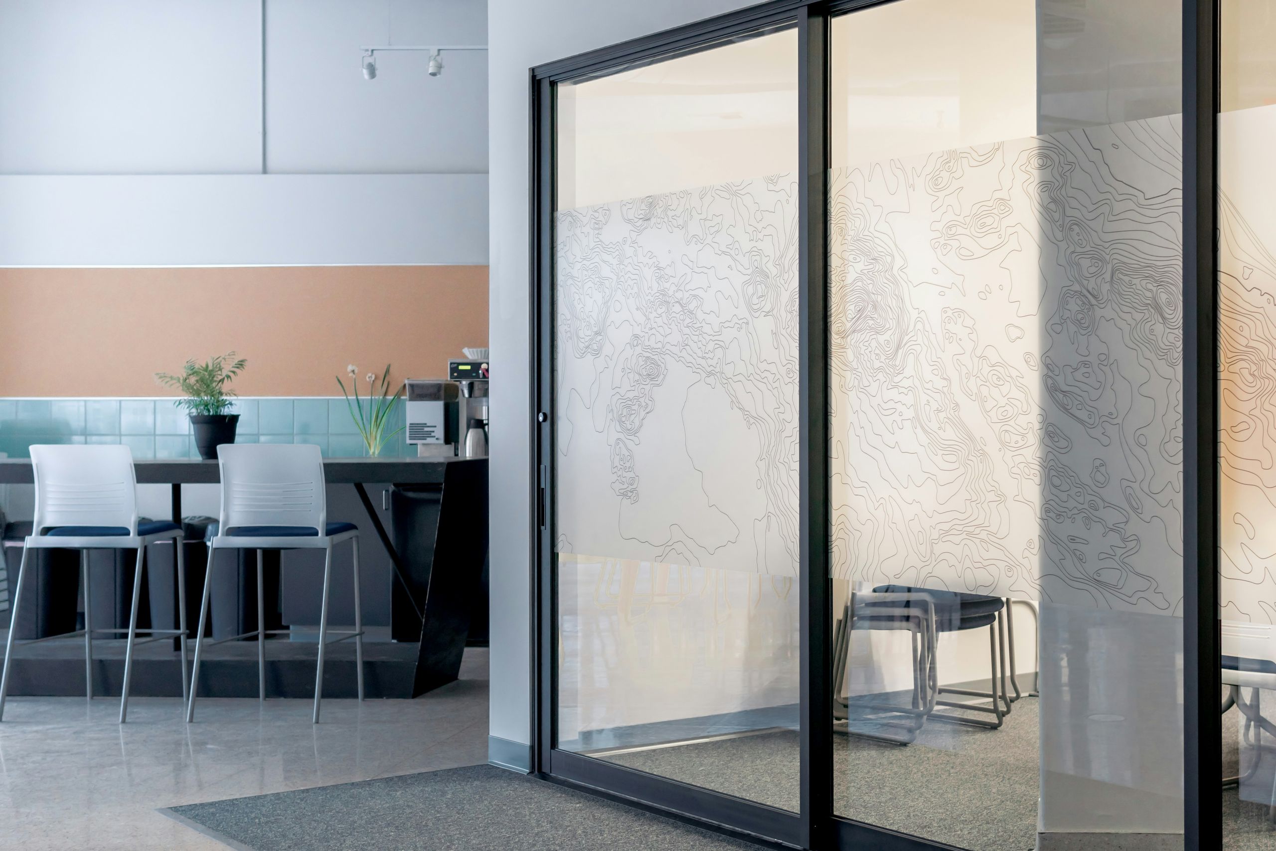 a glass door replacement showing a modern office space with glass sliding doors, a small meeting area with chairs behind patterned frosted glass, and a kitchenette with bar stools, a countertop, and plants in the background.