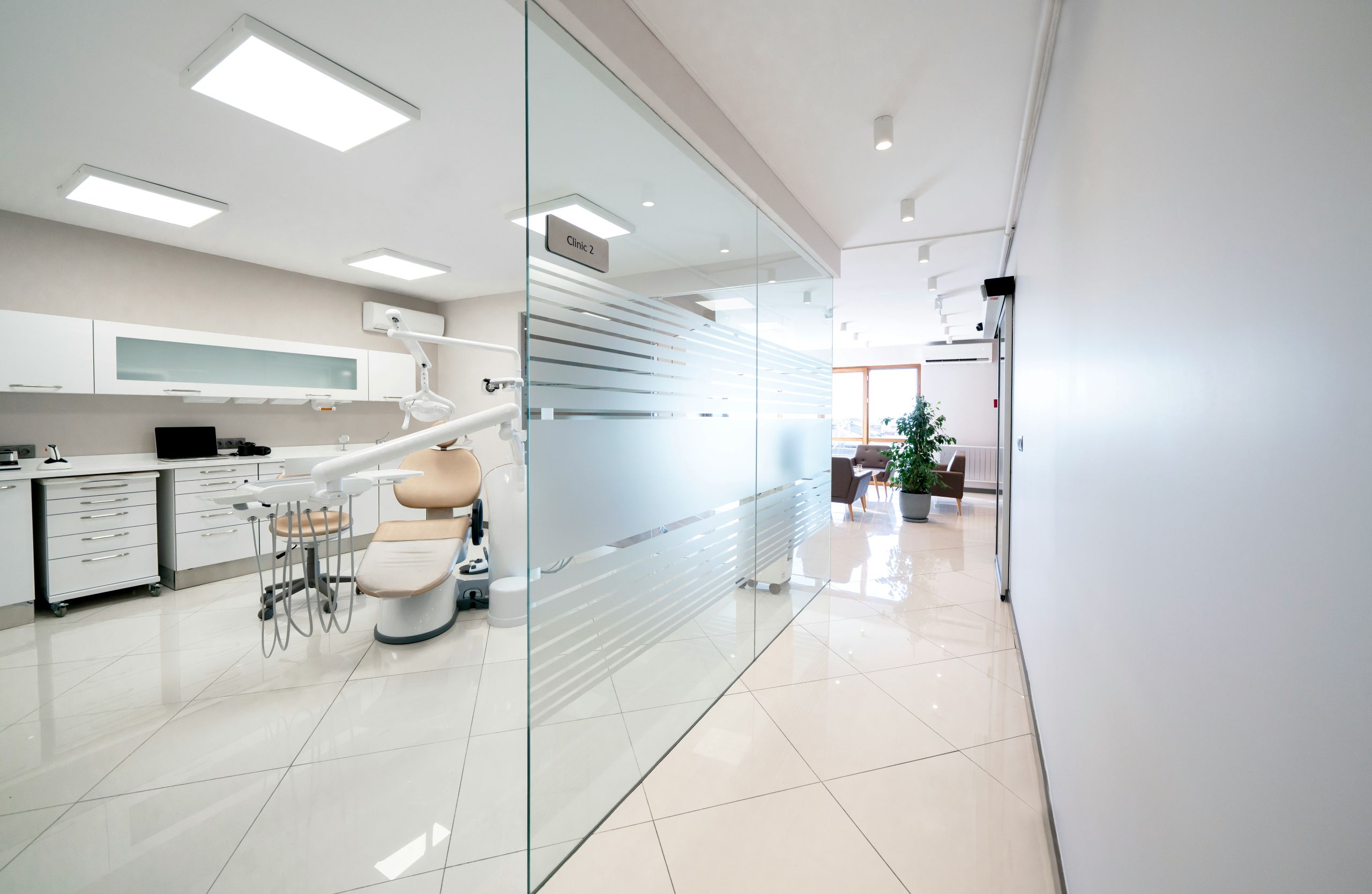 A commercial glass installation of modern dental clinic with white walls, a glass partition, dental chair, equipment, and a bright waiting area with plants visible through the glass.