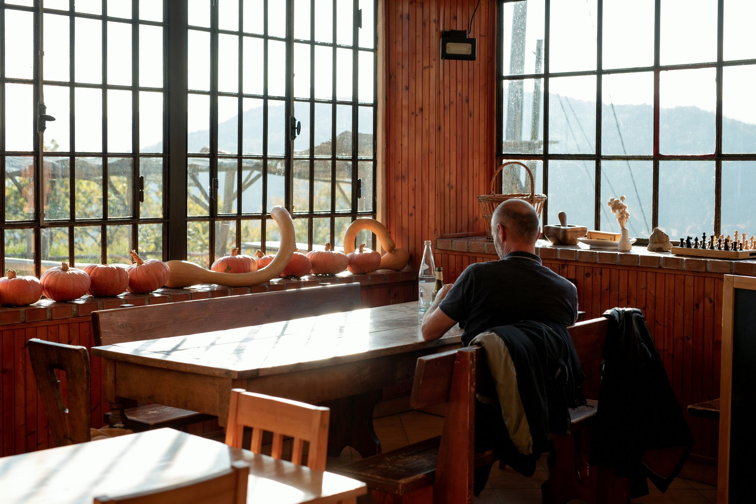 A person sits alone at a wooden table in a sunlit room with large windows, surrounded by pumpkins and gourds on the windowsill. Mountains are visible outside.